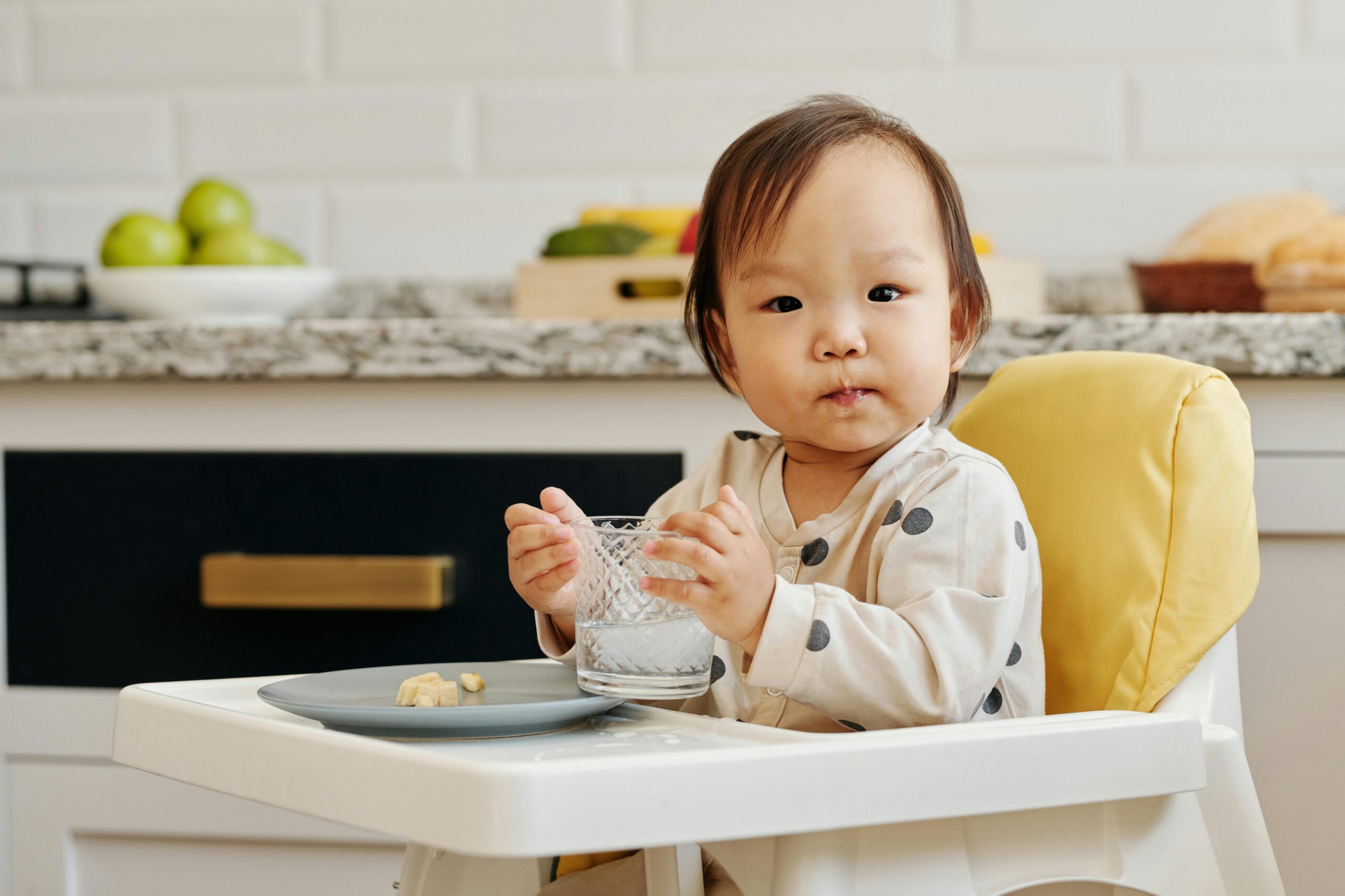 Adorable toddler sitting in a high chair, enjoying a meal and drink inside a cozy kitchen.